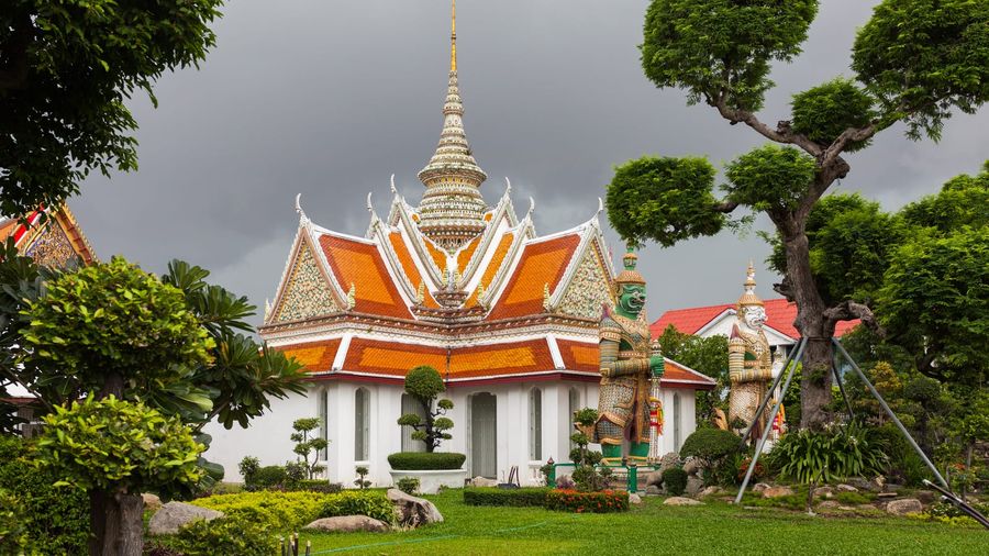 The white-and-orange tiled pavilion of Bangkok's Wat Arun temple crowned by an ornate spire, flanked by mythical guardian giant statues and manicured tropical gardens under a stormy grey sky
