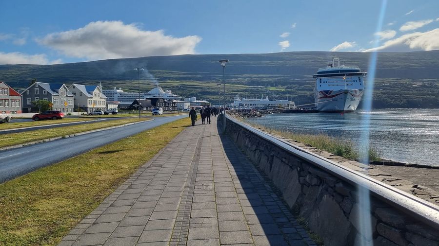 Pedestrians on Akureyri's harbourside promenade beside a road and timber houses, with a cruise ship docked in Eyjafjörður fjord and mountains rising behind
