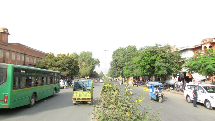 A green JCTSL city bus, a blue battery e-rickshaw, white taxis and motorbikes sharing a tree-lined main road beside the terracotta buildings of Jaipur's old city near Hawa Mahal