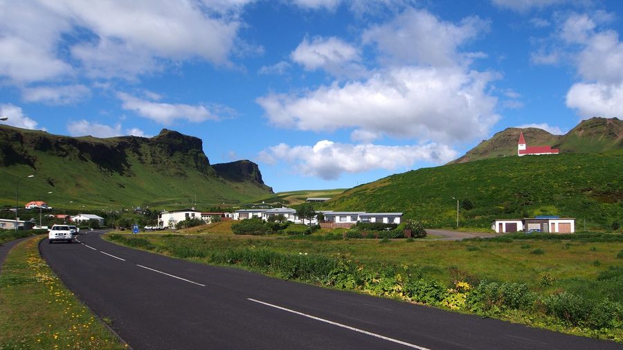 A car on the paved road into Vík í Mýrdal passing the village's white houses, with the red-roofed Víkurkirkja church on the green hillside and mountains rising beyond under a blue sky
