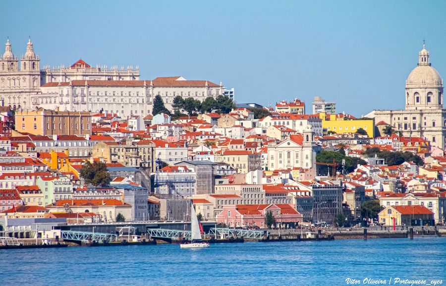Lisbon's pastel houses and terracotta roofs rising from the Tagus riverfront, with the white Monastery of Sao Vicente de Fora and the dome of the National Pantheon on the skyline and a sailboat in the foreground
