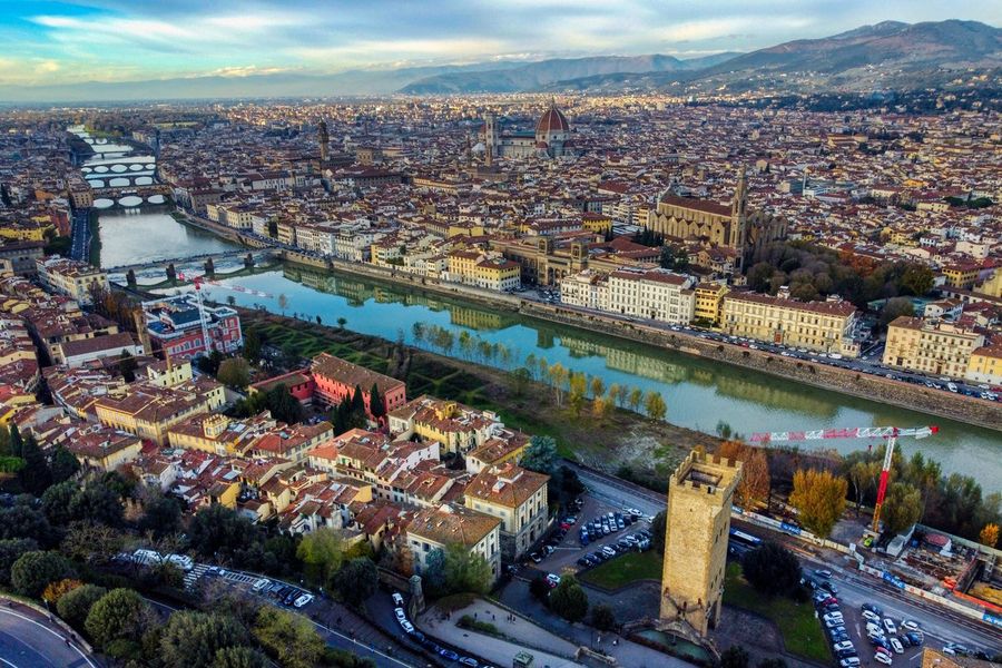 An aerial view of Florence along the river Arno, with the red dome of the Duomo, the tower of Palazzo Vecchio and the Basilica of Santa Croce among the terracotta rooftops and hills beyond