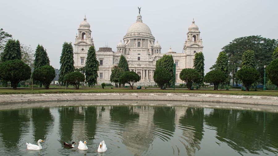 The white marble Victoria Memorial in Kolkata mirrored in its garden pond, with ducks on the water and manicured trees framing the domed facade under a hazy sky
