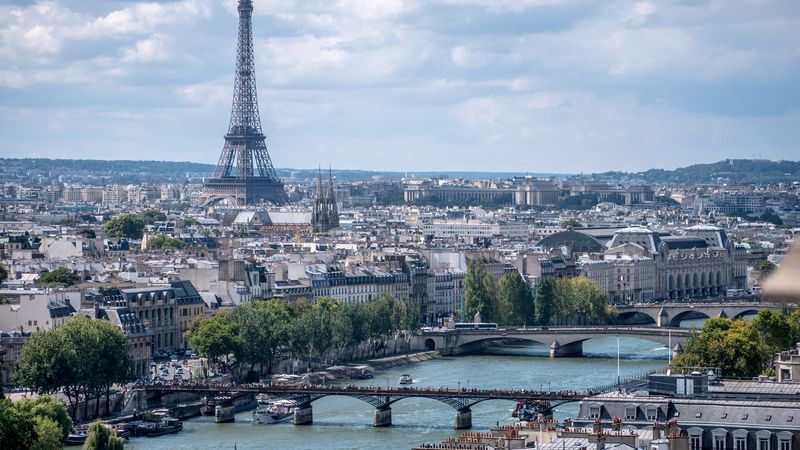 The Eiffel Tower rising above the rooftops of Paris with bridges spanning the River Seine in the foreground under a partly cloudy sky