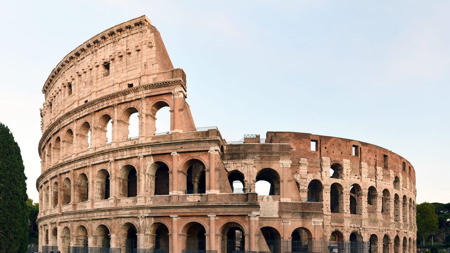 The tiered stone arches and weathered façade of the Roman Colosseum lit by warm evening light against a clear pale sky